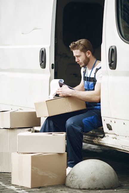 A young male mover with a beard, dressed in blue overalls and a white t-shirt, is sitting at the edge of a white van with its side door open. He is holding a clipboard and appears to be reviewing paperwork or inventory lists related to a house removal. Several cardboard boxes of various sizes are placed on the ground next to him, some stacked and some open, revealing packing materials inside. The scene takes place outdoors on a paved area with a concrete bollard nearby, suggesting a loading zone or driveway. The image captures the process of packing and organizing belongings for a home relocation, with visible furniture wrapping and packing supplies, typical of a professional removals service. The setting illustrates the loading process, highlighting the careful handling and documentation involved in furniture transport, as provided by Man With a Van Mile End.