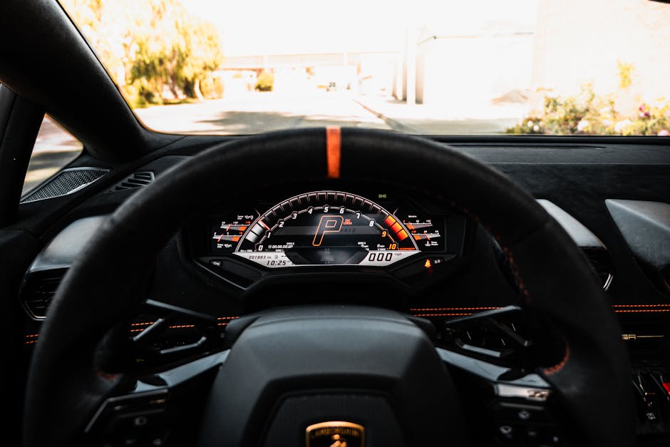 The image shows the interior of a modern car viewed from the driver's perspective, with the steering wheel prominently in the foreground. Behind the wheel, the digital dashboard displays a clean, illuminated interface with a large speedometer, tachometer, and other vehicle status indicators. The gear is in 'P' (park), and the odometer reads approximately 3,118 miles. The interior features a sleek dashboard with black and red accents, and the steering wheel has a red stripe at the top center. Through the windshield, a driveway or street with trees and a fence is visible under a clear, bright sky. The lighting inside the vehicle is ambient, highlighting the details on the dashboard and steering wheel. This scene relates to vehicle transport or packing during a home relocation process, possibly associated with a removal service such as Man With a Van Mile End, focusing on the transport aspect of house removals.
