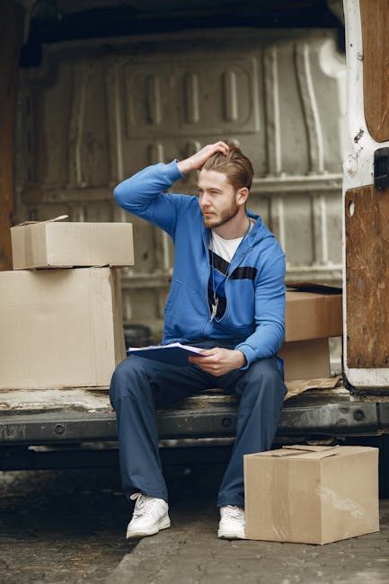 A young man with light brown hair wearing a blue hoodie, blue trousers, and white sneakers is sitting on the edge of the open back of a large moving van, surrounded by packed cardboard boxes and some smaller containers. He is holding a clipboard and appears to be contemplating or reviewing a checklist, with his left hand on his head. The interior of the van reveals a plain, light-colored, ribbed metal wall, and the back door is open, showing the pavement outside. Several cardboard boxes of various sizes are placed both inside and outside the van, some stacked and others resting on the ground, indicating an ongoing home relocation or furniture transport process. The scene is well-lit with natural light, emphasizing the logistics and organization involved in packing and preparing for a house move, as provided by Man With a Van Mile End.