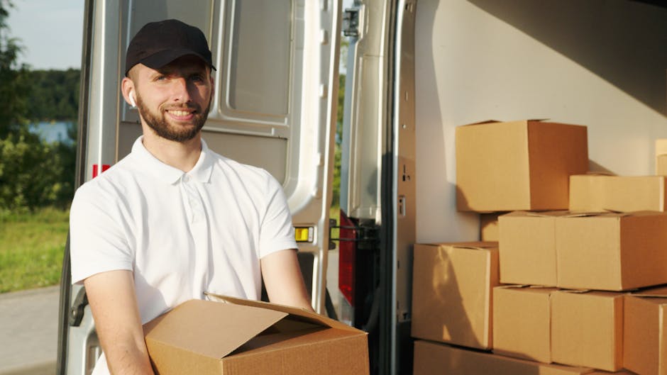 A male mover from Man With a Van Mile End stands outside a moving van, holding a medium-sized cardboard box during a home relocation process. The van's rear doors are open, revealing neatly stacked cardboard boxes of various sizes inside, some wrapped in plastic or packing paper to secure contents. The setting is outdoors on a paved driveway with greenery, trees, and a body of water visible in the background, indicating a suburban or rural location. The mover is dressed in a white polo shirt and black cap, smiling slightly as he prepares to load or unload additional boxes, with lifting and packing equipment such as trolleys, straps, or blankets possibly nearby. The natural lighting highlights the orderly packing and loading process typical of furniture transport and packing during house moves, reflecting professional removal services carried out by Man With a Van Mile End.
