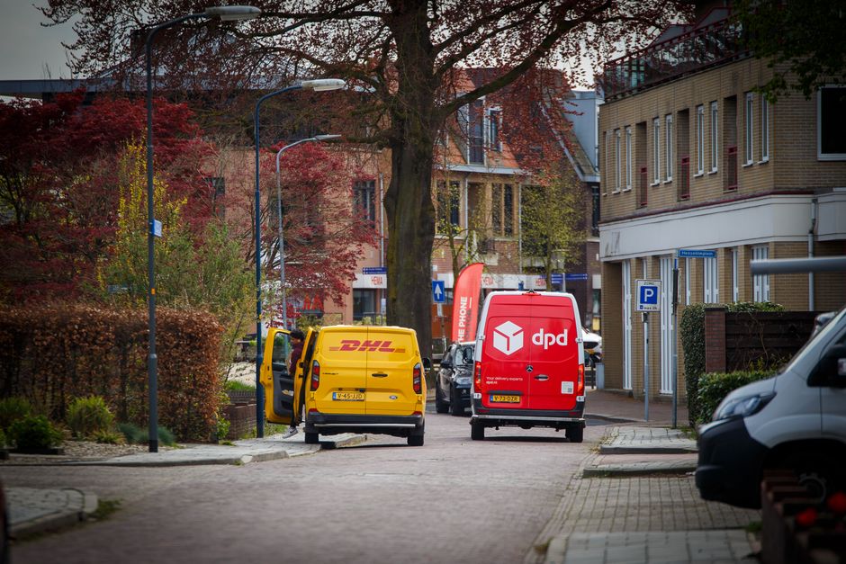 A white moving van parked on a street in front of a multi-storey residential building with grey stone facade. The van's rear doors are open, revealing a loading area filled with various packing materi
