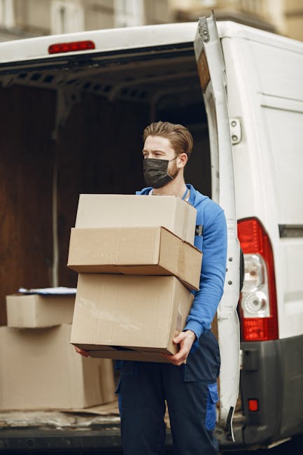 A man with light skin, wearing a black face mask and a blue jacket, is standing outside at the rear of a white commercial van with its back door open. He is holding three cardboard boxes, stacked one