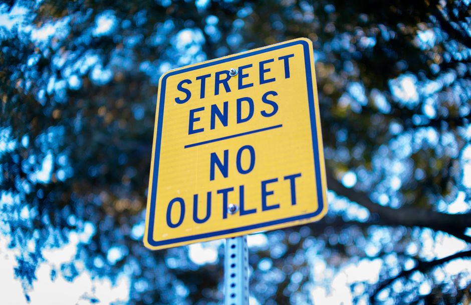 A close-up view of a yellow rectangular street sign with bold blue lettering reading 'STREET ENDS NO OUTLET,' mounted on a metal pole against a background of blurred dark green and blue foliage. The sign is situated outdoors, indicating the termination of a street without an outlet, which could impact house removals or transportation routes. The background lighting suggests daylight, and the sign is clearly visible, providing essential information for residents and removal service providers planning logistics and moving activities in the Mile End area, especially amid local road closures and van restrictions as referenced on the Mile End Park page. This image captures the signage that may influence parking and vehicle routes during home relocation tasks involving [COMPANY_NAME], supporting efficient planning for furniture transport and packing during moves.