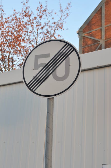 A round traffic sign displaying a 50 miles per hour speed limit with a black diagonal line crossing out the number, mounted on a metal pole. Behind the sign, there is a white corrugated metal fence and a brick building with a triangular gable visible in the upper right corner. In the background, there are bare tree branches and a clear blue sky. The scene suggests an urban environment in Mile End, with road restrictions potentially impacting home relocation or furniture transport services. Man With a Van Mile End may occasionally encounter such signage during local house removals and moving logistics, requiring careful planning for loading and unloading processes in areas with road closures or restrictions, including Mile End Park road closures and van restrictions.