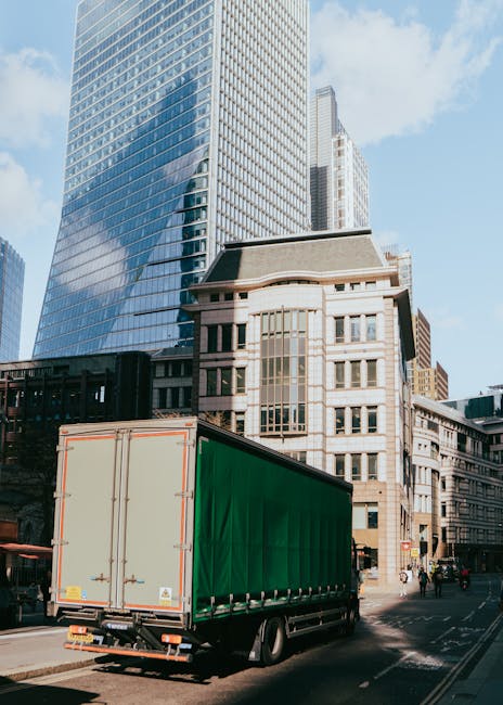 A large green box truck with closed rear doors is positioned on a city street during daylight hours, adjacent to modern high-rise office buildings and older stone structures. The truck is part of a home relocation or furniture transport operation, with the vehicle's cargo area visible and the street showing typical urban surroundings, including pedestrians and a paved roadway. The scene is well-lit by natural sunlight, with clear blue skies overhead. This image illustrates the loading process involved in professional removals services provided by companies like Man With a Van Mile End, supporting efficient packing and moving logistics in an urban environment.