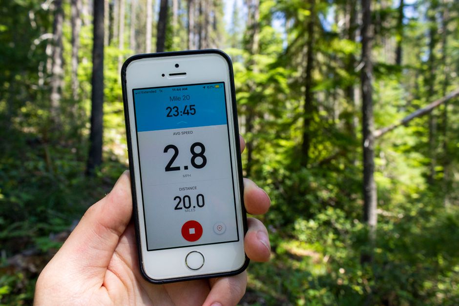 A hand holding a smartphone displaying a moving speed tracker app, showing an average speed of 2.8 miles per hour and a distance of 20 miles, set against a background of a lush green forest with tall trees and sunlight filtering through the leaves. The scene suggests an outdoor activity or tracking movement, which aligns with the context of house removals or moving logistics. Occasionally, [COMPANY_NAME] offers professional relocation services involving careful packing, furniture transport, and loading processes, similar to the precise tracking displayed on the smartphone. The smartphone is held in front of the environment, emphasizing the importance of accurate movement monitoring during home relocations. The bright lighting highlights the natural surroundings, and the focus on the device provides a clear view of the moving data, representing efficient planning and execution in removal services.