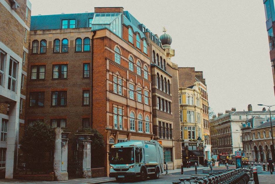 A large green box truck with closed rear doors is positioned on a city street during daylight hours, adjacent to modern high-rise office buildings and older stone structures. The truck is part of a home relocation or furniture transport operation, with the vehicle's cargo area visible and the street showing typical urban surroundings, including pedestrians and a paved roadway. The scene is well-lit by natural sunlight, with clear blue skies overhead. This image illustrates the loading process involved in professional removals services provided by companies like Man With a Van Mile End, supporting efficient packing and moving logistics in an urban environment.