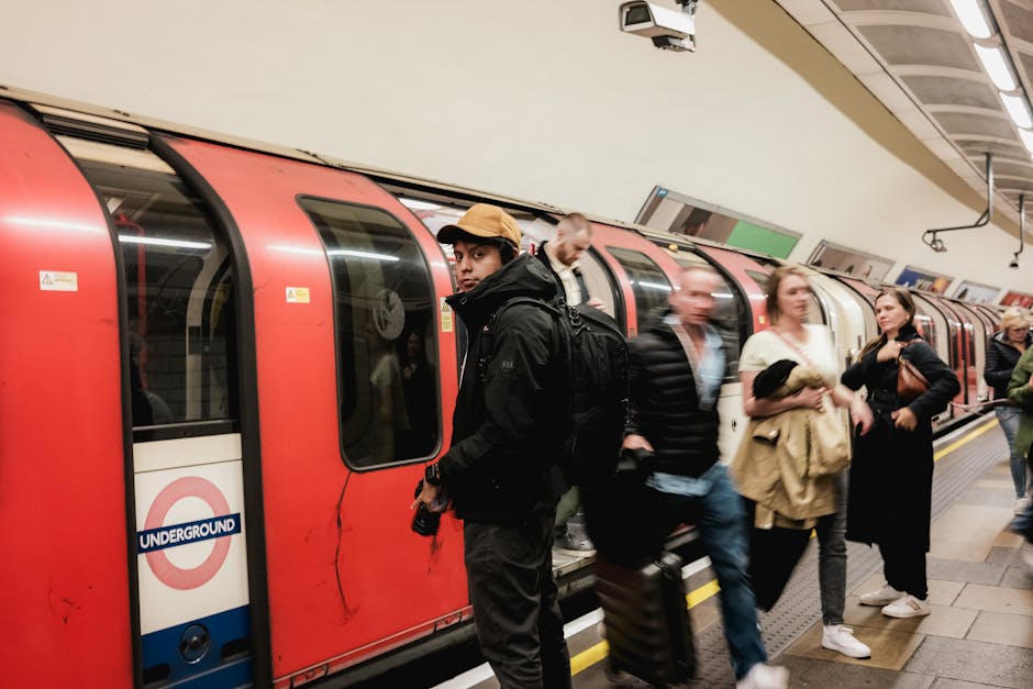 Inside a London Underground station, several commuters are standing on the platform near a red train with sliding doors and windows, some holding luggage and bags. A young man wearing a black jacket, backpack, and beige cap is looking towards the camera, while others, including a woman in a beige coat and a man in a striped jacket, are engaged in conversation or preparing to board. The platform has a tiled floor and a low yellow safety line along the edge. Overhead, there are ceiling-mounted lights and signs, with a monitor displaying travel information. The scene captures a typical busy underground environment, relevant to the context of house removals and home relocation logistics, as shown in services provided by Man With a Van Mile End, with personnel often assisting customers with packing or moving plans near transport hubs like Mile End Tube Station.