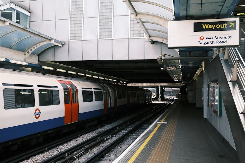 Inside a London Underground station, several commuters are standing on the platform near a red train with sliding doors and windows, some holding luggage and bags. A young man wearing a black jacket, backpack, and beige cap is looking towards the camera, while others, including a woman in a beige coat and a man in a striped jacket, are engaged in conversation or preparing to board. The platform has a tiled floor and a low yellow safety line along the edge. Overhead, there are ceiling-mounted lights and signs, with a monitor displaying travel information. The scene captures a typical busy underground environment, relevant to the context of house removals and home relocation logistics, as shown in services provided by Man With a Van Mile End, with personnel often assisting customers with packing or moving plans near transport hubs like Mile End Tube Station.