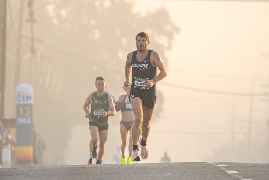 A group of male runners participating in a road race during early morning or late afternoon, with soft, warm sunlight illuminating their figures. The lead runner is wearing a black athletic singlet with the word 'Illinois' and 'Elite' printed on it, along with black shorts and running shoes. Behind him, two other male runners in sleeveless tops and running shorts are visible, both focused on the race, with one slightly behind and another further back. The runners are on an asphalt road, and in the background, there are blurred trees, power lines, and a race marker post indicating mile 13. The asphalt appears slightly elevated, and the runners are captured mid-stride, demonstrating an active, dynamic scene typical of a competitive or organized race event. The lighting creates a slightly hazy, atmospheric effect, emphasizing the movement and determination of the runners, which subtly links to the themes of logistics and transportation associated with house removals and moving services provided by Man With a Van Mile End.