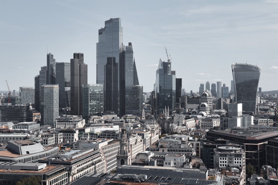 A cityscape view of central London with numerous modern skyscrapers, including tall glass office buildings with reflective surfaces and cranes on top indicating ongoing construction. In the foreground, there are older, shorter buildings with flat rooftops, some with rooftop installations and drainage systems. The scene is captured during daylight hours with a partly cloudy sky illuminating the scene. This urban environment showcases the high-rise skyline typical of a busy business district, which could relate to professional office removals services such as those provided by Man With a Van Mile End, especially when considering logistics involving furniture or office equipment transfer during a corporate relocation or packing and moving process.