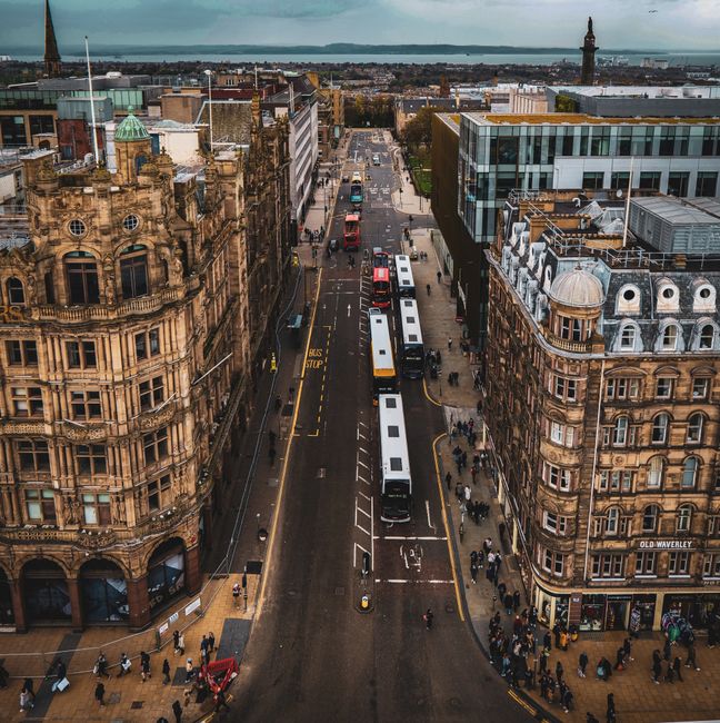 An aerial view of a busy city street in Mile End with several white and black buses parked along the curb, surrounded by ornate historical buildings with decorative facades and large windows. The street is filled with pedestrians walking on the sidewalks, some carrying bags, while others wait at bus stops or cross the road. The scene includes a clear, cloudy sky and a distant view of the river with ships and industrial structures on the horizon. The environment reflects typical urban activity, with signage indicating bus stops and street markings visible on the asphalt. The image captures the transportation and urban landscape associated with home relocation or moving services like those offered by Man With a Van Mile End, illustrating the logistics involved in furniture transport and packing during a house move.