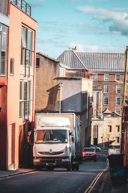 A white delivery van parked on a narrow urban street surrounded by multi-storey residential and commercial buildings with brick and stucco facades, some featuring large windows and balconies. The van is positioned close to a building on the left side, with its rear facing the camera, indicating the loading process typical of home relocation or furniture transport. A black trash bin is visible on the right pavement, and a few other vehicles, including a red car, are parked further down the street. The sky above is partly cloudy, casting shadows on the buildings, illustrating a typical day for a local moving service such as Man With a Van Mile End, which specializes in house removals and packing and moving in the Mile End, Victoria Park, and Bow areas.