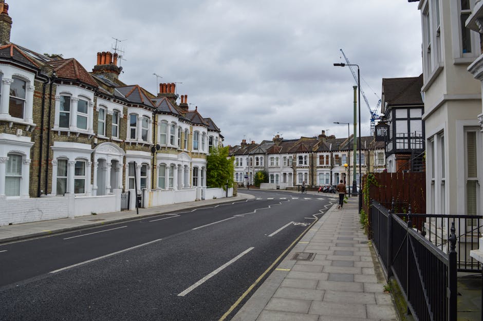 A quiet residential street in Mile End during daytime with several Victorian-style terraced houses featuring white facades, bay windows, and red-tile roofs. A grey pavement runs alongside the row of houses, separated from the asphalt road by a black metal fence. The road has white dashed lane markings and is currently empty of vehicles, except for a white van parked in the distance. Several street lamps line the pavement, and a person is walking along the sidewalk. Overcast weather with grey clouds creates diffuse lighting that highlights the architectural details of the houses. This street scene relates to house removals and relocation services, illustrating an urban environment where furniture transport and home moving logistics could occur, with Man With a Van Mile End potentially operating in similar settings.