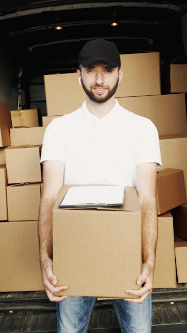 A young man with a beard, wearing a black cap, white polo shirt, and blue jeans, is standing at the open rear of a black van during a home relocation. He is holding a medium-sized cardboard box in front of him, with a clipboard resting on top of it. Behind him, the van's interior is filled with stacked cardboard boxes of various sizes, some wrapped in plastic or placed directly on the vehicle floor. To the left, larger boxes are visible, arranged in a way that indicates they are ready for loading or unloading. The scene is outdoors with natural lighting, capturing the process of packing, packing materials, and furniture transport as part of the moving logistics handled by Man With a Van Mile End, supporting professional removals near Queen Mary University Mile End E3.