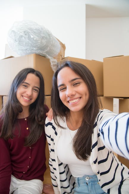 Two young women with smiling expressions are taking a selfie in a home interior surrounded by moving boxes. The woman on the right, wearing a white shirt and a striped jacket, is holding the camera and leaning slightly forward, capturing her and her friend in the frame. The woman on the left, dressed in a maroon top, stands close with her arm around her friend's shoulder. Behind them, several large cardboard boxes are stacked, some with bubble wrap covering objects placed on top of the boxes. The setting appears to be a room prepared for moving, with natural lighting illuminating the scene. This moving process, managed by Man With a Van Mile End, exemplifies home relocation tasks such as packing, loading, and transportation of household items, highlighting the importance of careful handling during furniture transport and the packing and moving stages of a house removal near Queen Mary University Mile End E3.