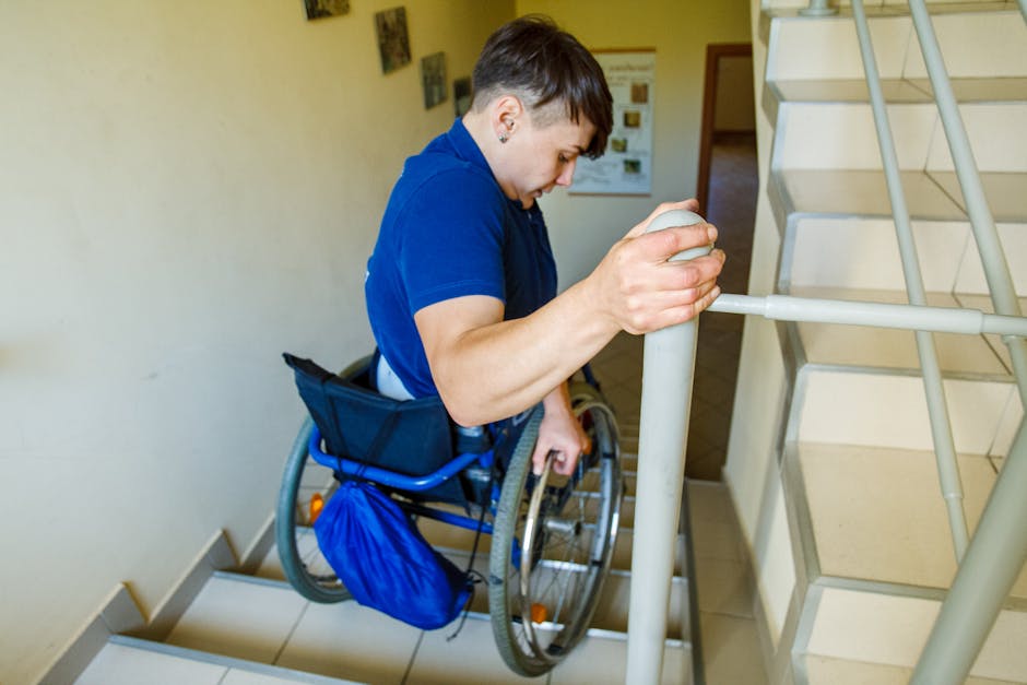 A person in a wheelchair with short dark hair wearing a blue shirt, holding the handrail of a staircase with their right hand while positioned on a staircase inside a residential building. The individual appears to be preparing for or engaged in a moving process as part of a home relocation or furniture transport. The scene shows the staircase with white tiled steps, a beige wall, and a metal handrail. The person's wheelchair features black fabric and orange safety reflectors, with a blue bag attached to the side. The interior environment is well-lit, and there are some framed pictures or documents visible on the wall in the background. This image exemplifies the challenges faced in stairs and parking when performing removals, highlighting the need for professional services like those provided by Man With a Van Mile End, especially for accessible moving and packing procedures.