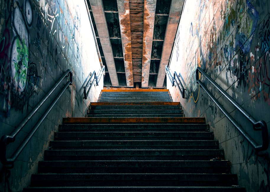 A set of dark, weathered concrete stairs with orange safety strips along the edge of each step, leading upward inside an underpass or tunnel with graffiti-covered walls on both sides. The walls are painted with various tags and artwork in colors including green, blue, and black. Steel handrails are fixed along both sides of the staircase for support. The ceiling above the stairs features exposed wooden beams and metal supports, with a small amount of natural light filtering in from the top of the stairs. This environment may be part of a residential or commercial building entryway, requiring careful navigation during a home relocation or furniture transport process, as managed by Man With a Van Mile End, specialists in removals and moving logistics.