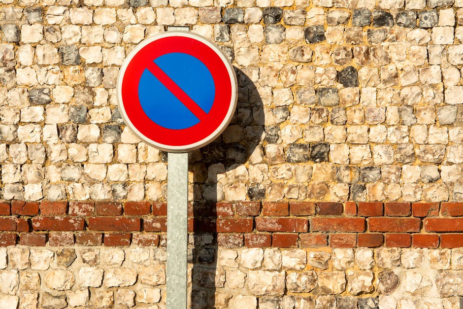 A round no parking sign featuring a red border, blue background, and a red diagonal line across the center is mounted on a metal pole and positioned in front of a textured brick wall composed of light-colored stones interspersed with darker stones. The shadow of the sign falls onto the brick wall, indicating sunlight from the left side. The setting suggests a street or urban area where parking restrictions are enforced, relevant to home relocation and furniture transport processes. Such signage is important for planning vehicle placement during house removals in Mile End, as managed by Man With a Van Mile End, particularly under local parking permits rules outlined for Tower Hamlets removals.