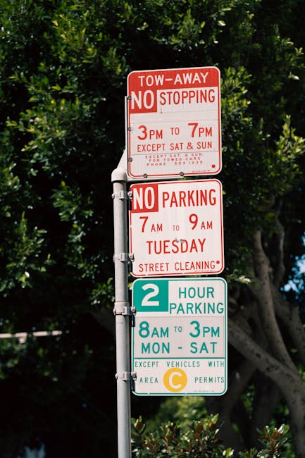 A multi-level parking and parking restriction sign mounted on a grey metal pole outdoors, with a background of green leafy trees and blue sky. The top sign, in red and white, indicates tow-away zones and no stopping from 3 pm to 7 pm, with additional details about parking restrictions and street cleaning. Below it, another red and white sign designates no parking from 7 am to 9 am on Tuesdays for street cleaning. The bottom sign, in blue and white, permits 2-hour parking from 8 am to 3 pm, Monday through Saturday, with a note that vehicles displaying a C area permit are exempt. The signs are effectively used by Man With a Van Mile End for managing parking regulations in relation to local house removals and furniture transport services in the Mile End area.