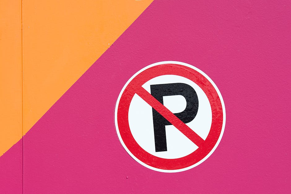 A round no parking sign featuring a red border, blue background, and a red diagonal line across the center is mounted on a metal pole and positioned in front of a textured brick wall composed of light-colored stones interspersed with darker stones. The shadow of the sign falls onto the brick wall, indicating sunlight from the left side. The setting suggests a street or urban area where parking restrictions are enforced, relevant to home relocation and furniture transport processes. Such signage is important for planning vehicle placement during house removals in Mile End, as managed by Man With a Van Mile End, particularly under local parking permits rules outlined for Tower Hamlets removals.
